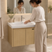 Bassino 600 mm wall-mounted bathroom vanity with glossy white ceramic basin, natural oak PVC cabinet, stainless-steel support and two-door storage.