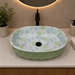 Green blue and white floral ceramic table top wash basin with glossy finish, oval designer countertop basin measuring 500 × 320 × 140 mm for modern bathrooms.